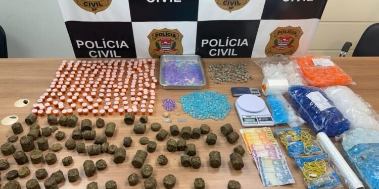 Mesa com drogas apreendidas pela DEIC Piracicaba, incluindo maconha, cocaína, dinheiro e materiais para embalo, em frente a painel da Polícia Civil de São Paulo.