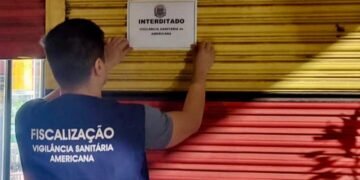 Agente da Vigilância Sanitária de Americana fixa aviso de interdição em porta metálica de estabelecimento durante operação de fiscalização noturna.