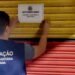 Agente da Vigilância Sanitária de Americana fixa aviso de interdição em porta metálica de estabelecimento durante operação de fiscalização noturna.