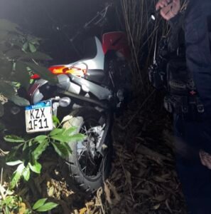 Motocicleta vermelha roubada, localizada pela ROMU Piracicaba em área de mata durante a noite, com um guarda civil iluminando o local com lanterna.
