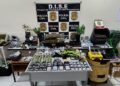 Laboratório de maconha gourmet desmontado pela DISE com drogas, estufas e materiais apreendidos expostos sobre mesas. #PraCegoVer: A imagem mostra uma sala com diversas drogas organizadas sobre mesas, incluindo porções embaladas, tabletes e plantas semelhantes à maconha. Ao fundo há um painel da Polícia Civil com identificação da DISE. Também aparecem balanças, celulares, embalagens e equipamentos utilizados no preparo e armazenamento das drogas.