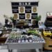 Laboratório de maconha gourmet desmontado pela DISE com drogas, estufas e materiais apreendidos expostos sobre mesas. #PraCegoVer: A imagem mostra uma sala com diversas drogas organizadas sobre mesas, incluindo porções embaladas, tabletes e plantas semelhantes à maconha. Ao fundo há um painel da Polícia Civil com identificação da DISE. Também aparecem balanças, celulares, embalagens e equipamentos utilizados no preparo e armazenamento das drogas.