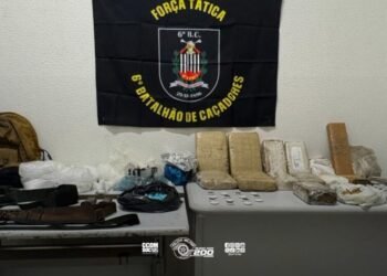 Mais de 20 quilos de drogas apreendidos pela Força Tática organizados sobre mesas com acessórios e carregadores. #PraCegoVer: A imagem mostra uma sala com duas mesas onde estão expostos tabletes e porções de drogas embaladas, além de carregadores de arma, cintos táticos e equipamentos. Ao fundo há uma bandeira da Força Tática do 6º Batalhão.