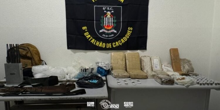 Mais de 20 quilos de drogas apreendidos pela Força Tática organizados sobre mesas com acessórios e carregadores. #PraCegoVer: A imagem mostra uma sala com duas mesas onde estão expostos tabletes e porções de drogas embaladas, além de carregadores de arma, cintos táticos e equipamentos. Ao fundo há uma bandeira da Força Tática do 6º Batalhão.