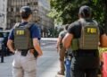 Apreensão de 40 gramas de metanfetamina após investigação do FBI com agentes em patrulhamento urbano. #PraCegoVer: A imagem mostra dois agentes com coletes identificados com a sigla FBI nas costas caminhando por uma calçada em área urbana. Ao fundo aparecem prédios comerciais, uma viatura policial estacionada e árvores ao lado da via.