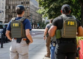 Apreensão de 40 gramas de metanfetamina após investigação do FBI com agentes em patrulhamento urbano. #PraCegoVer: A imagem mostra dois agentes com coletes identificados com a sigla FBI nas costas caminhando por uma calçada em área urbana. Ao fundo aparecem prédios comerciais, uma viatura policial estacionada e árvores ao lado da via.