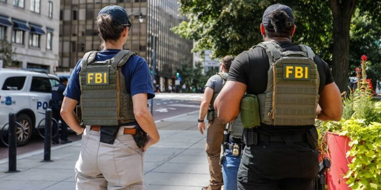 Apreensão de 40 gramas de metanfetamina após investigação do FBI com agentes em patrulhamento urbano. #PraCegoVer: A imagem mostra dois agentes com coletes identificados com a sigla FBI nas costas caminhando por uma calçada em área urbana. Ao fundo aparecem prédios comerciais, uma viatura policial estacionada e árvores ao lado da via.