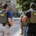 Apreensão de 40 gramas de metanfetamina após investigação do FBI com agentes em patrulhamento urbano. #PraCegoVer: A imagem mostra dois agentes com coletes identificados com a sigla FBI nas costas caminhando por uma calçada em área urbana. Ao fundo aparecem prédios comerciais, uma viatura policial estacionada e árvores ao lado da via.