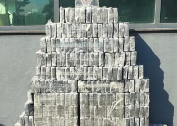 300 quilos de cocaína apreendidos pelo TOR organizados em tabletes empilhados. #PraCegoVer: A imagem mostra diversos tabletes retangulares de droga embalados em plástico transparente, empilhados em formato de pirâmide em frente a uma parede com janela de vidro. No topo da pilha há um brasão policial.