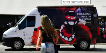 como denunciar assédio moral e sexual no trabalho #PraCegoVer mulher virada de costas com cabelo loiro, blusa preta e calça jeans, indo sentido a uma van com uma mulher desenhada e uma boca maior escrito "Precisamos falar do assédio