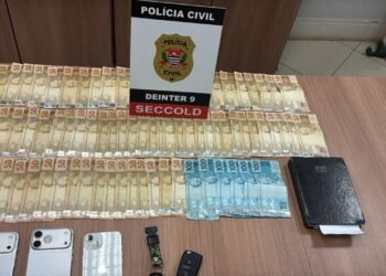 operação tiger iii contra lavagem de dinheiro #PraCegoVer: sobre uma mesa estão diversas notas de dinheiro organizadas em fileiras, aparelhos celulares, uma chave de veículo, um relógio e um caderno, ao fundo um painel da Polícia Civil com identificação do DEINTER 9 e SECCOLD