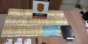 operação tiger iii contra lavagem de dinheiro #PraCegoVer: sobre uma mesa estão diversas notas de dinheiro organizadas em fileiras, aparelhos celulares, uma chave de veículo, um relógio e um caderno, ao fundo um painel da Polícia Civil com identificação do DEINTER 9 e SECCOLD