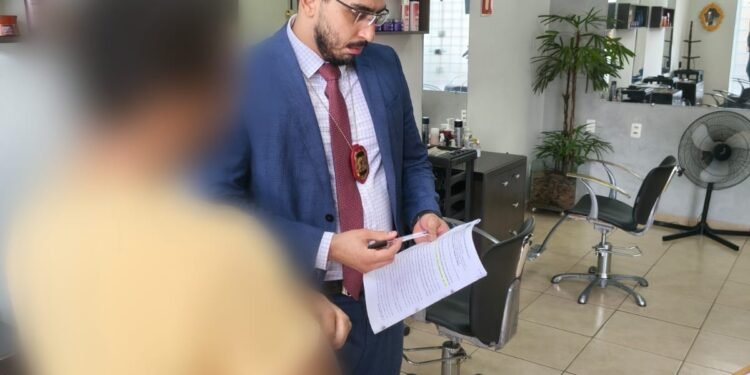 polícia civil investiga ameaças contra primeira-dama do df #PraCegoVer: policial civil em trajes formais segura documentos durante cumprimento de mandado em um salão comercial com cadeiras e espelhos ao fundo