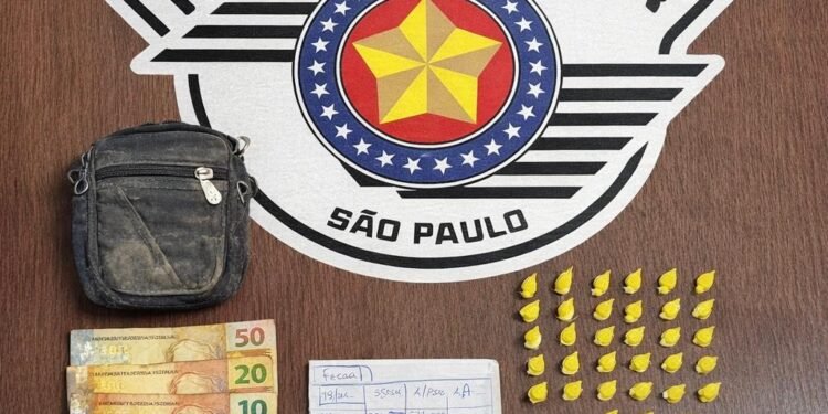 Apreensão de tráfico de drogas realizada pela Polícia Militar em Leme #PraCegoVer Na imagem aparecem 33 eppendorfs amarelos com cocaína organizados sobre uma mesa, dinheiro em notas de real, um celular, uma pequena bolsa e anotações do tráfico ao fundo com o brasão da Polícia Militar de São Paulo.