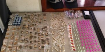 guarda municipal apreende drogas em condomínio bom sucesso 02 #PraCegoVer mesa com grande quantidade de porções de drogas embaladas, pinos coloridos, celulares e bonés da guarda municipal ao fundo