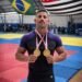 policial militar conquista ouro duplo no jiu jitsu apmbb 2026 #PraCegoVer: policial militar posa em tatame segurando duas medalhas de ouro no peito, com bandeiras do Brasil e do Estado de São Paulo ao fundo, durante campeonato de jiu-jítsu.