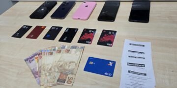 DIG prende trio por fraude contra idosos #PraCegoVer celulares, cartões bancários, dinheiro em espécie e comprovantes de atendimento bancário aparecem sobre uma mesa após apreensão feita pela Polícia Civil em ocorrência de fraude contra idosos