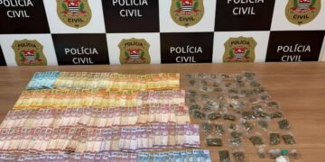 ROMU prende traficante na Comunidade Portelinha #PraCegoVer imagem mostra dinheiro em notas diversas, porções de drogas embaladas, frascos plásticos e uma folha com anotações sobre uma mesa, em frente a um painel da Polícia Civil de São Paulo.