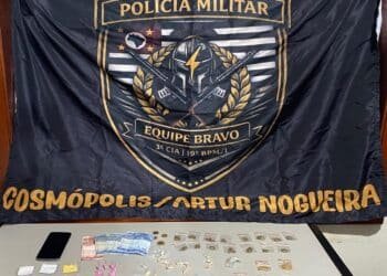 PM prende homem por trafico de drogas #PraCegoVer imagem mostra drogas, dinheiro e um celular apreendidos sobre uma mesa, com bandeira da Polícia Militar ao fundo