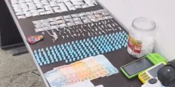 PM prende casal por tráfico de drogas em adega #PraCegoVer imagem mostra uma mesa com várias porções de drogas embaladas, microtubos, dinheiro, máquinas de cartão e câmeras de vigilância apreendidos pela Polícia Militar