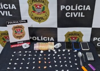 operação Open Doors III contra o tráfico de drogas #PraCegoVer: imagem mostra materiais apreendidos sobre uma mesa, com porções de drogas embaladas, dinheiro em espécie, três celulares, balança de precisão, colher, faca e isqueiros, diante de um painel da Polícia Civil de São Paulo e placa da DISE de Votuporanga.