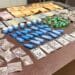 tráfico de drogas no Vida Nova #PraCegoVer: imagem mostra uma mesa com porções de drogas embaladas, dinheiro em notas e moedas, além de embalagens separadas para apresentação da ocorrência policial.